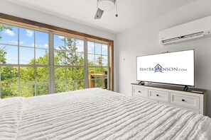 Ein Schlafzimmer mit einem großen Bett, einem Fernseher auf einer Kommode und einem Fenster mit Blick auf Bäume.