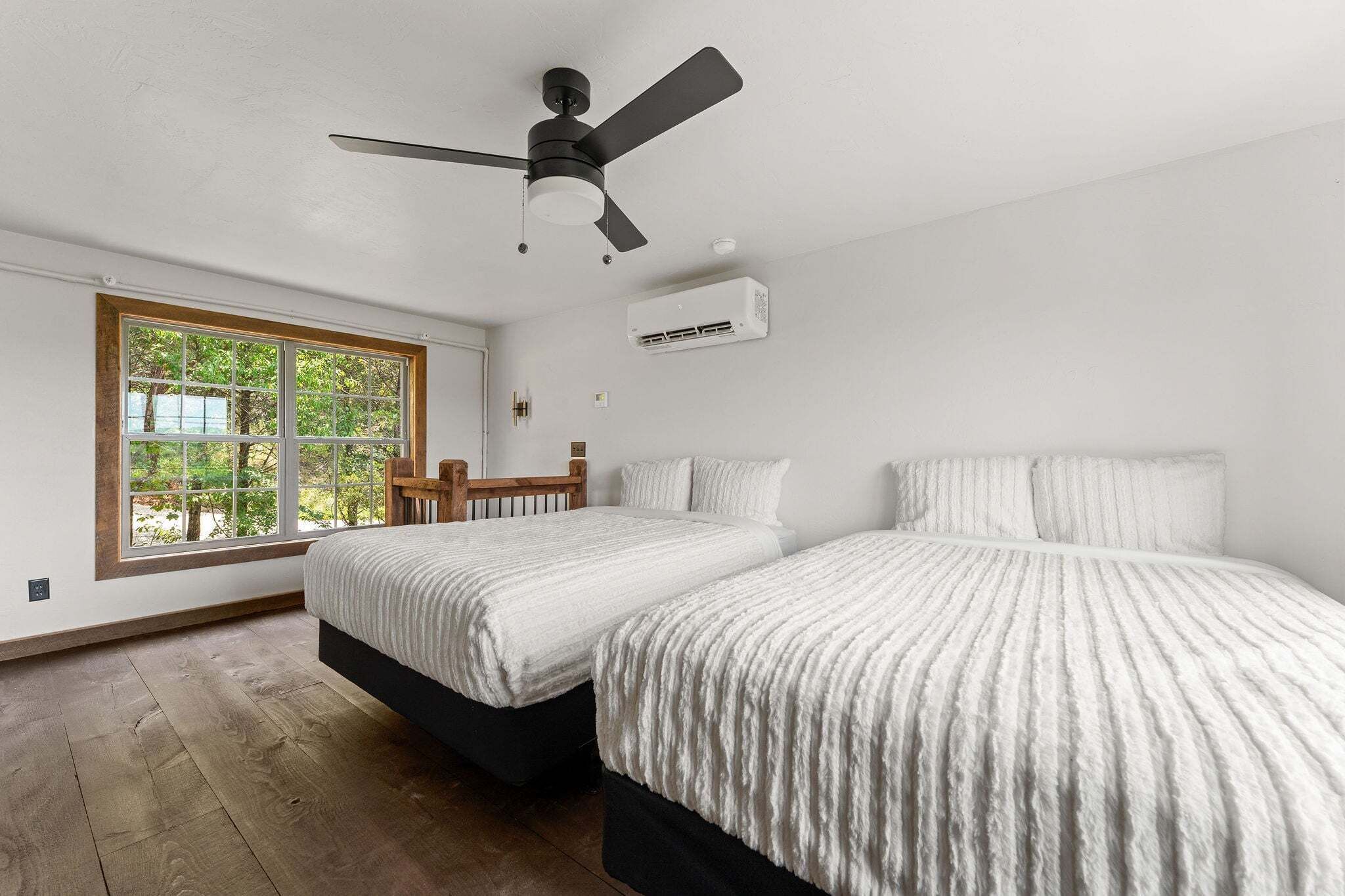 A bedroom with two beds, a ceiling fan, an air conditioning unit, and a window with a view of trees.
