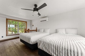 Ein Schlafzimmer mit zwei Betten, einem Deckenventilator, einer Klimaanlage und einem Fenster mit Blick auf Bäume.