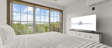 A bedroom with a large window, a bed with a textured blanket, a TV on a dresser, and an air conditioning unit on the wall.