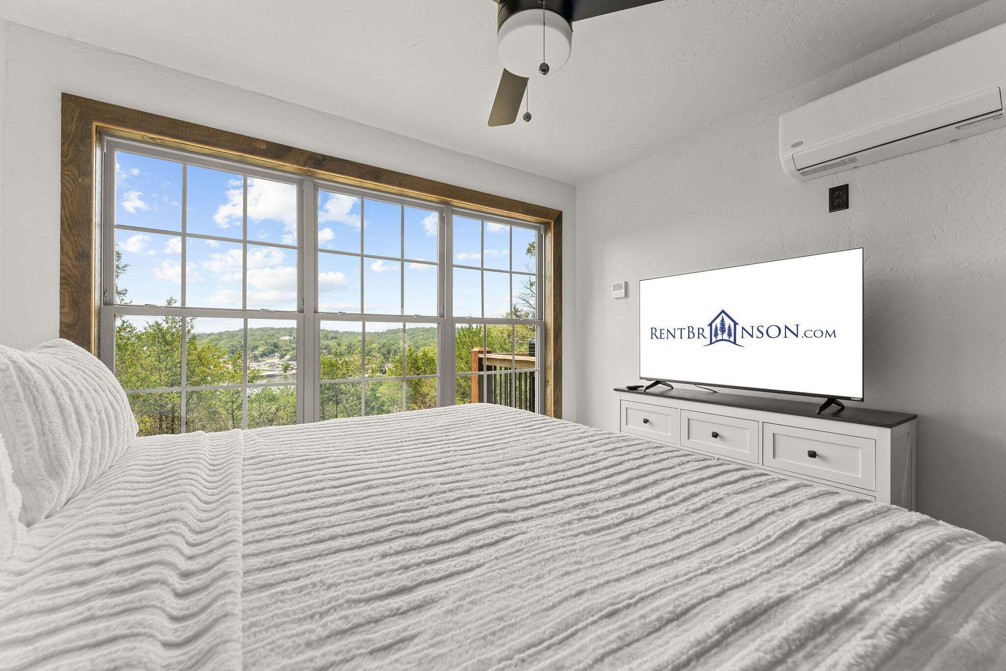 Un dormitorio con un ventanal grande, una cama con manta texturizada, un televisor de pantalla plana y un equipo de aire acondicionado en la pared.