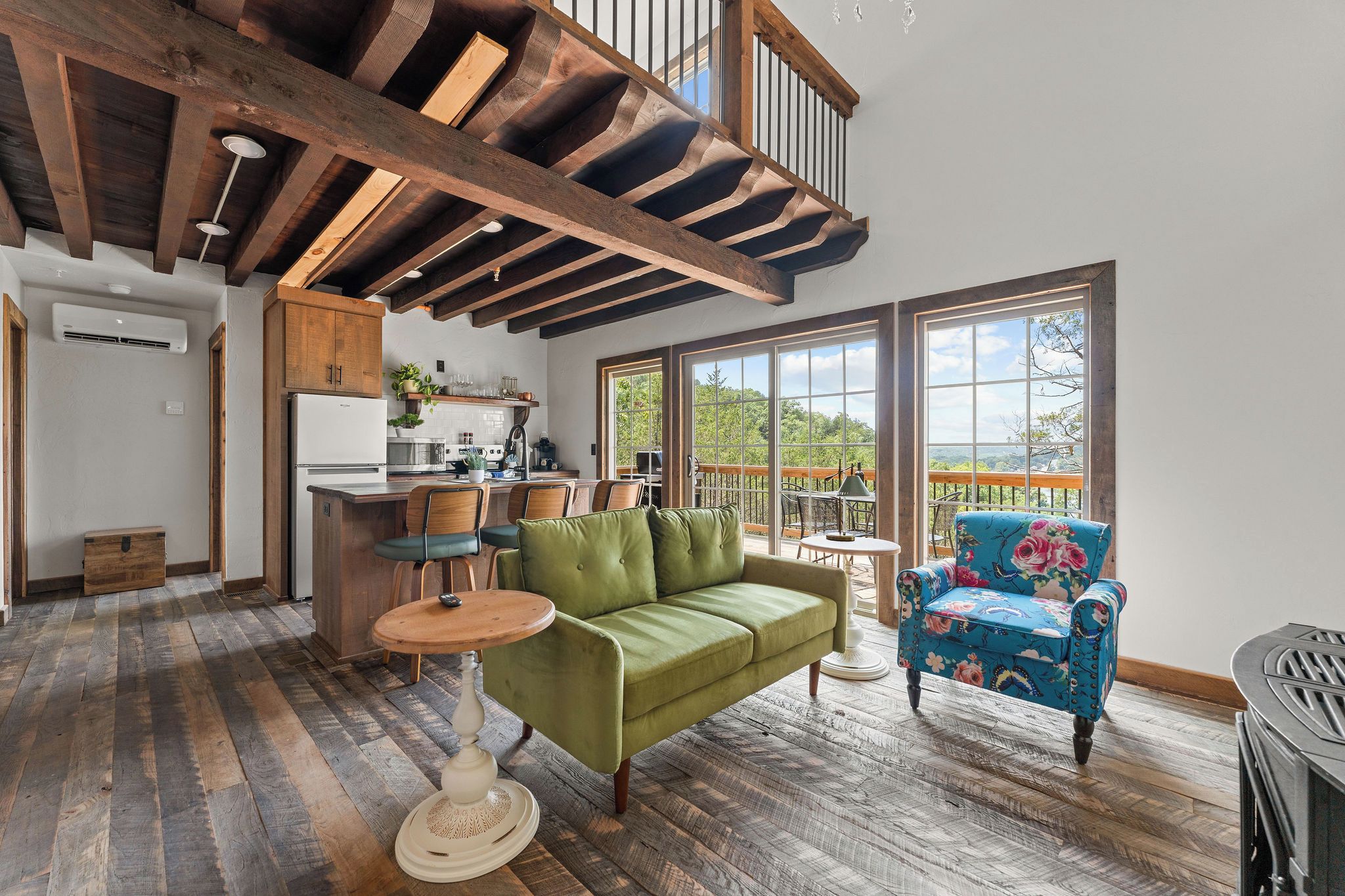 Uma sala de estar ampla com teto alto, vigas de madeira, um sofá verde, uma poltrona azul com padrão floral e vista através de grandes janelas.