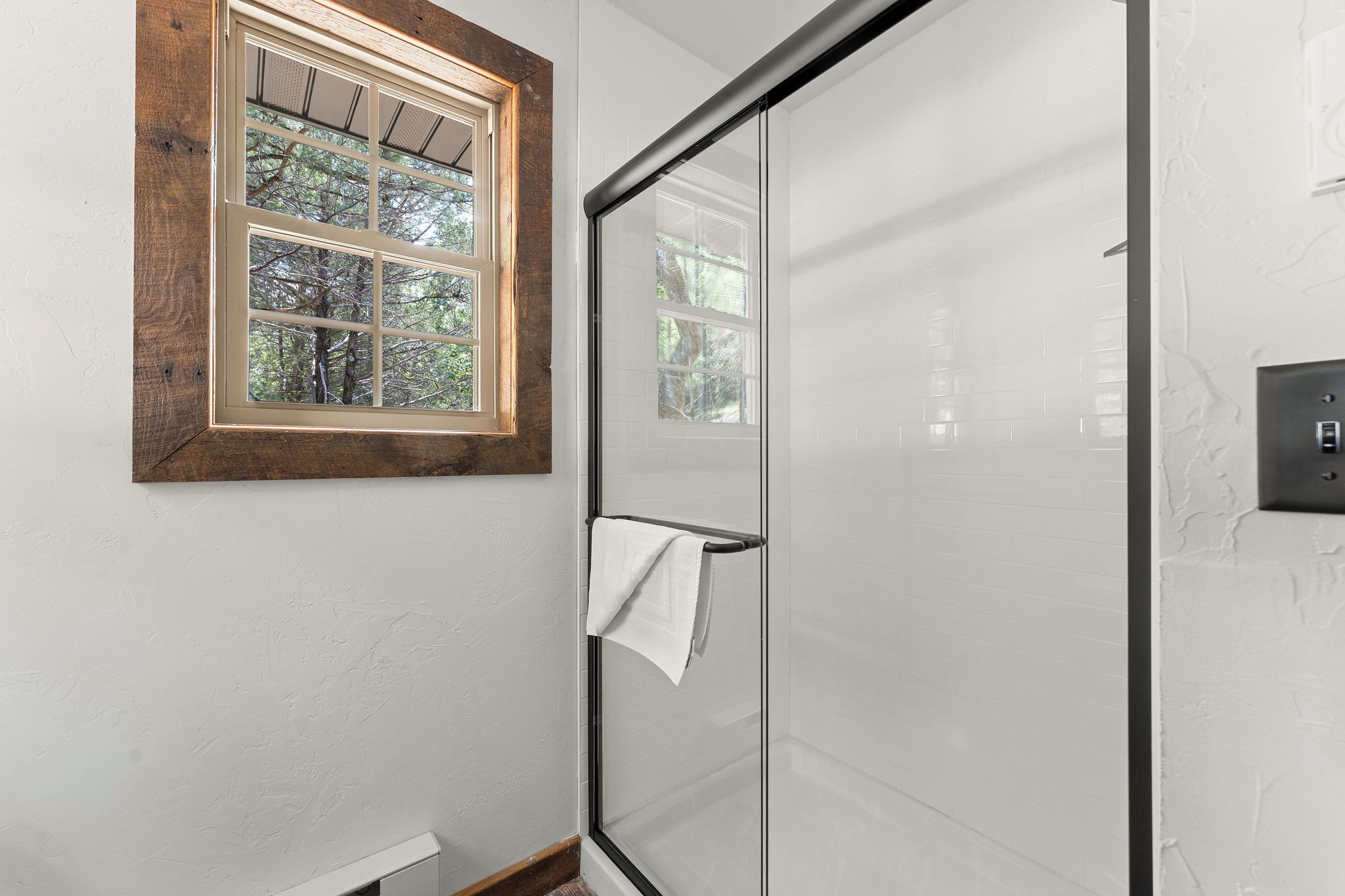 A bathroom with a glass shower door, a wooden-framed window, and a towel hanging on the door.