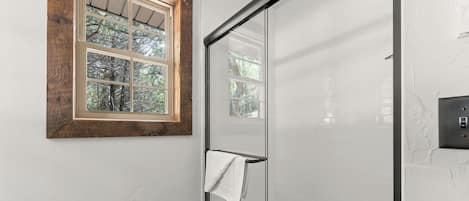 A bathroom with a glass shower door, a wooden-framed window, and a towel hanging on the door.