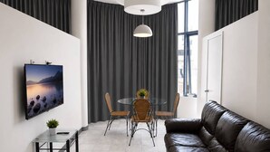 Un salon moderne avec un canapé en cuir, une table à manger avec des chaises, un téléviseur à écran plat fixé au mur et une grande fenêtre avec des rideaux.