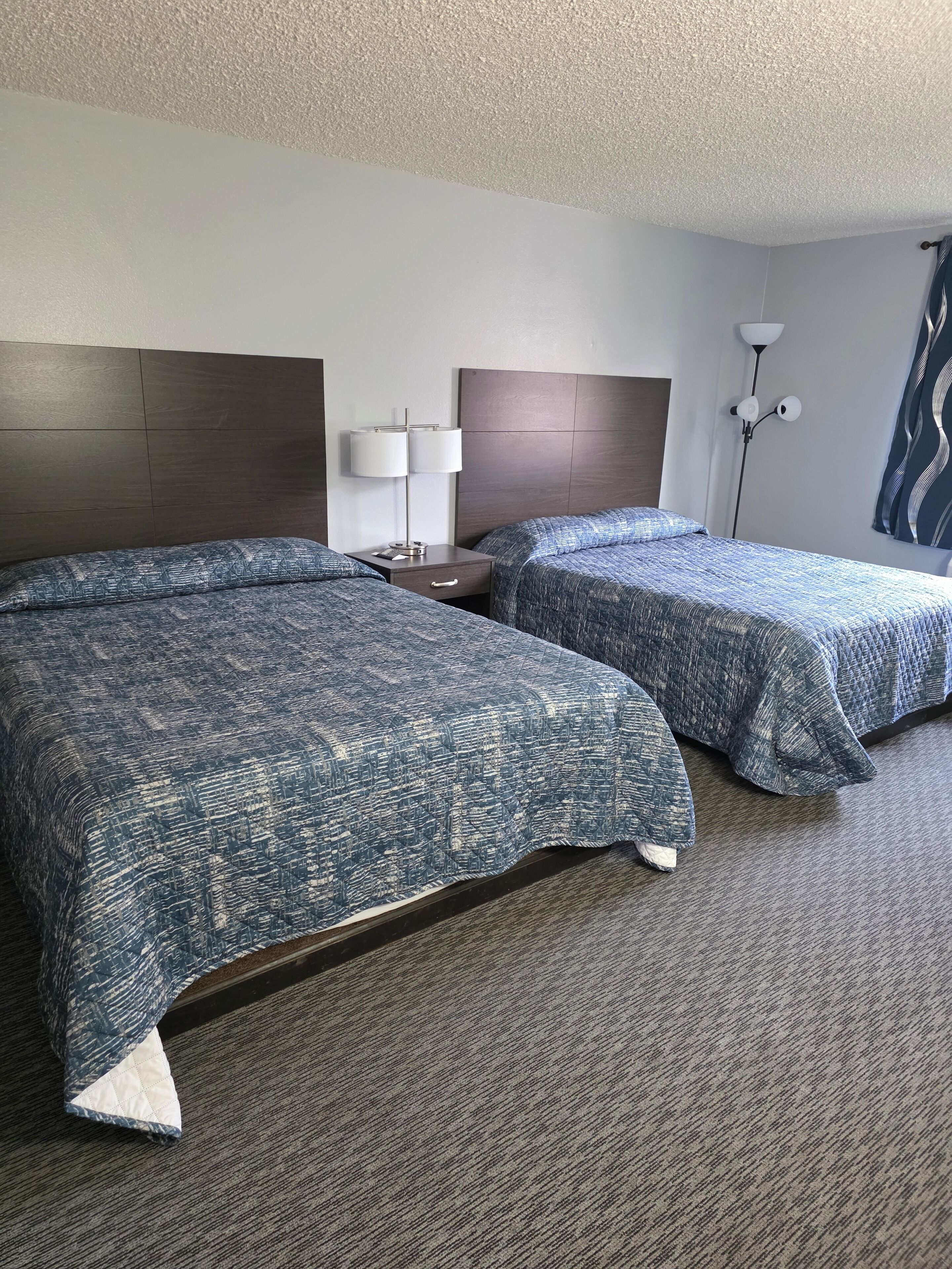 A hotel room with two beds, a nightstand, a lamp, and a wall-mounted light fixture.