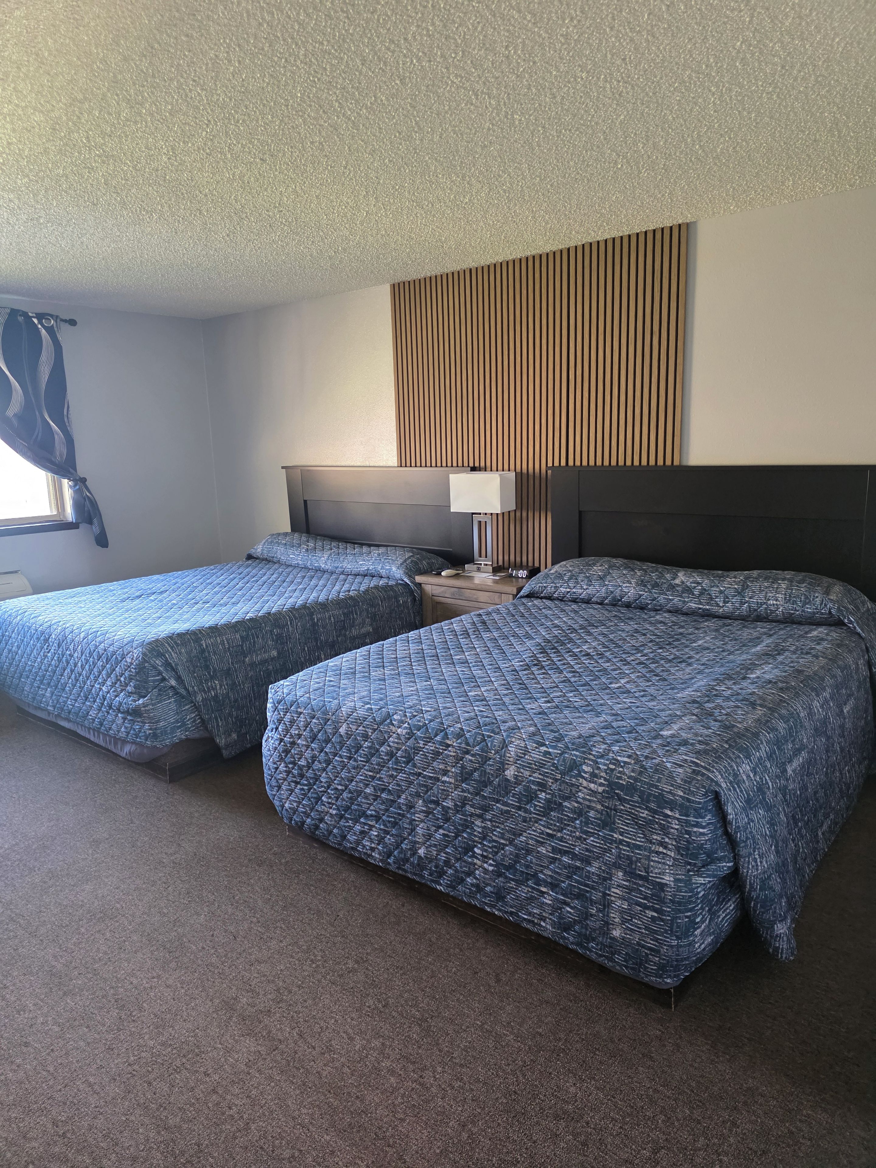 A hotel room with two beds, a nightstand, a lamp, and a window with curtains.
