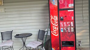 Coca-Colan myyntiautomaatti rakennuksen ulkopuolella, sen vieressä metallinen pöytä ja kaksi tuolia.
