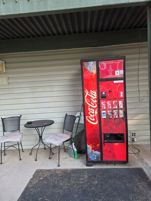 A Coca-Cola vending machine outside a building with a metal table and two chairs.