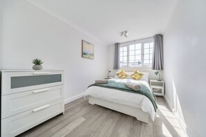 Ein Schlafzimmer mit Bett, Nachttischen, einem Schminktisch, einem Fenster mit Vorhängen und einer Deckenleuchte.