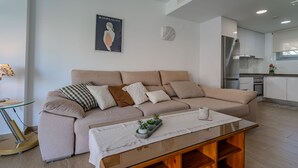 Un salon moderne avec un canapé beige, une table basse en verre et une cuisine en arrière-plan.