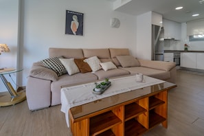 A modern living room with a beige sofa, a glass coffee table, and a kitchen in the background. - Villa Amalia 9: Ground floor with garden (Torrevieja)