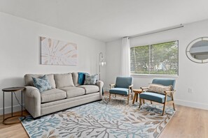Ein modernes Wohnzimmer mit einem grauen Sofa, zwei blauen Sesseln, einem gemusterten Teppich und einem großen Fenster mit Jalousien.