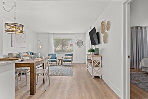 A modern living room with a dining area, a television mounted on the wall, and a window with blinds. - CLM 16B  Gorgeous, pet friendly - Private beach access (Destin)