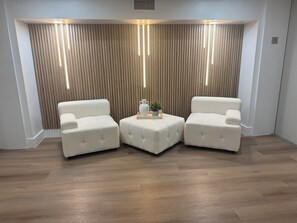 A room with three white tufted chairs arranged in a semi-circle around a central ottoman, which holds a vase and a small plant. The room features a wooden slatted wall with integrated lighting and a wooden floor. - Pristine one bedroom studio in downtown Fort Myers (Fort Myers)