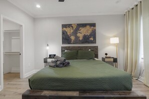 A bedroom with a large bed, a bedside table with a lamp, a wall-mounted artwork, and a door leading to another room.