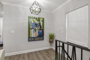 Ein Flur mit einer geometrisch gestalteten Deckenleuchte, einem Wandbild mit einer Szene vom Mardi Gras, einer Topfpflanze und einem Fenster mit Jalousien.