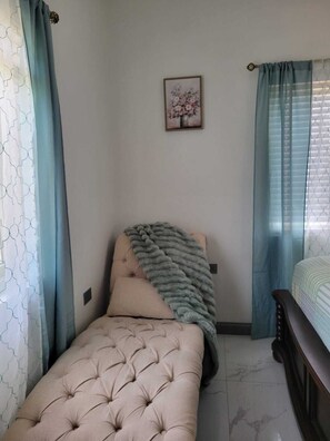 A bedroom with a single bed, a tufted headboard, a decorative pillow, a blanket, a framed floral artwork on the wall, and a window with curtains. - 3 Bedroom Luxe Hideaway in Ocho Rios with Pool Access (Ocho Rios)