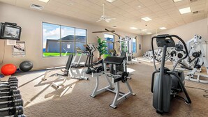 Ein Fitnessraum mit Trainingsgeräten, großen Fenstern und einem Deckenventilator.
