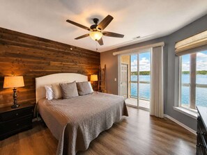 A bedroom with a wooden headboard, a bed with a textured blanket, a ceiling fan, and a view of a body of water through large windows. - Eagles Nest @ Lands End - Boat Slip! (Osage Beach)