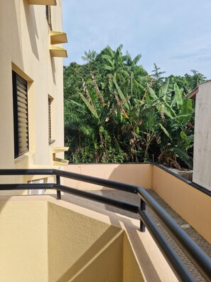 A balcony with a railing, a building with a window with blinds, and a lush green plant with large leaves. - Apto Confortável em Ubatuba a 4 Min da Praia das Toninhas com Wi-Fi e Garagem (Ubatuba)