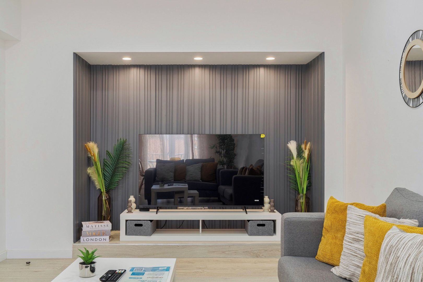 Ein modernes Wohnzimmer mit einem Flachbildfernseher, grauen Vorhängen und einem grauen Sofa mit gelben Kissen.