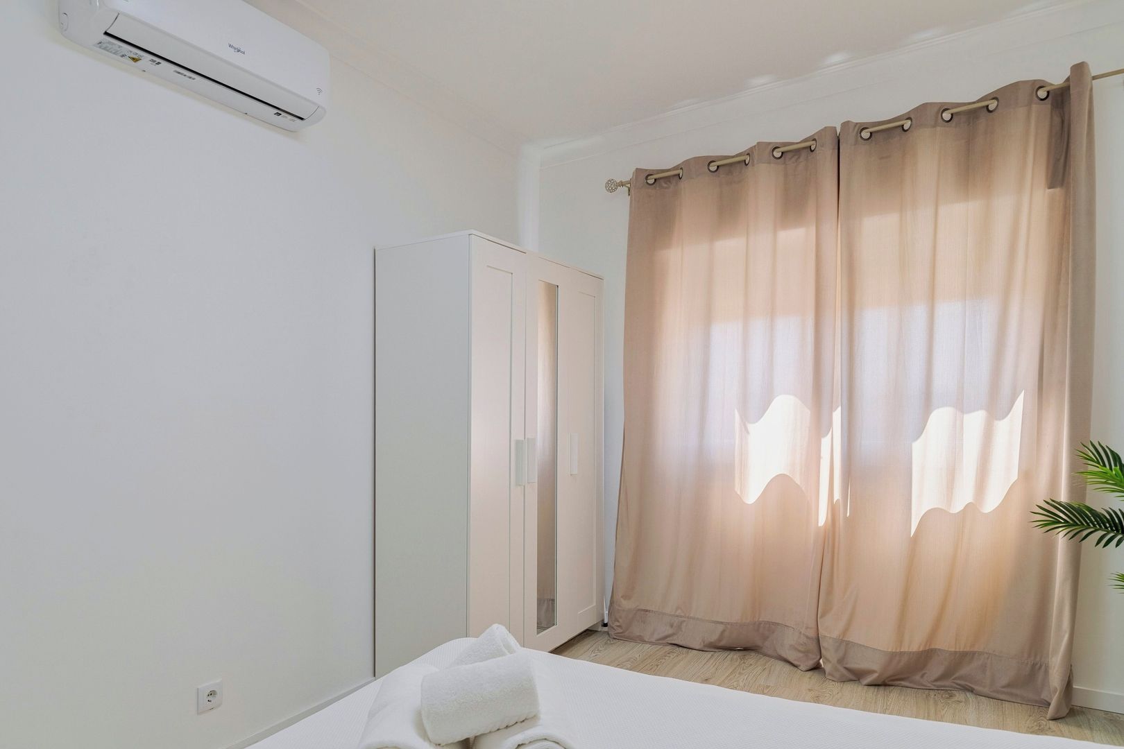A hotel room with a bed, a wardrobe, a window with sheer curtains, and an air conditioning unit.