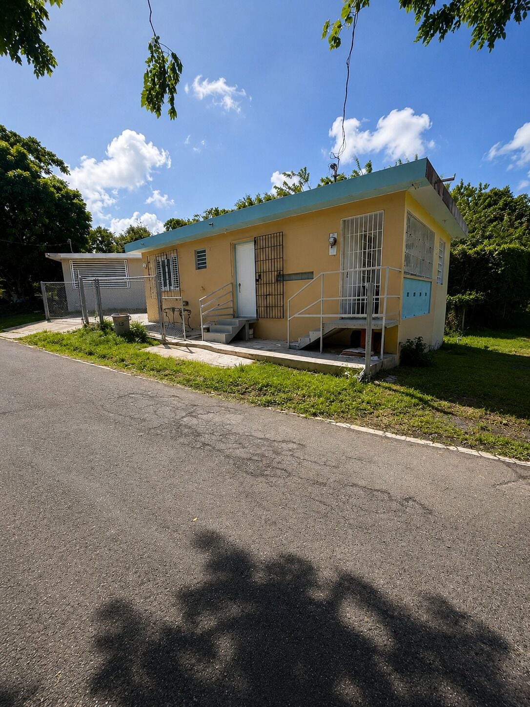 A small, single-story building with a flat roof, a door, and barred windows, situated next to a paved road with a grassy area in front.