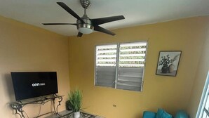 A living room with a sofa, a television, a ceiling fan, and a window with blinds. - The Naguabeña House of the Anvil (Ceiba)