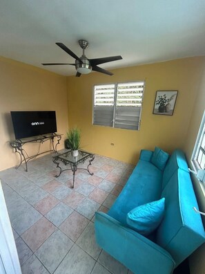 A living room with a sofa, a television, a ceiling fan, and a window with blinds. - The Naguabeña House of the Anvil (Ceiba)