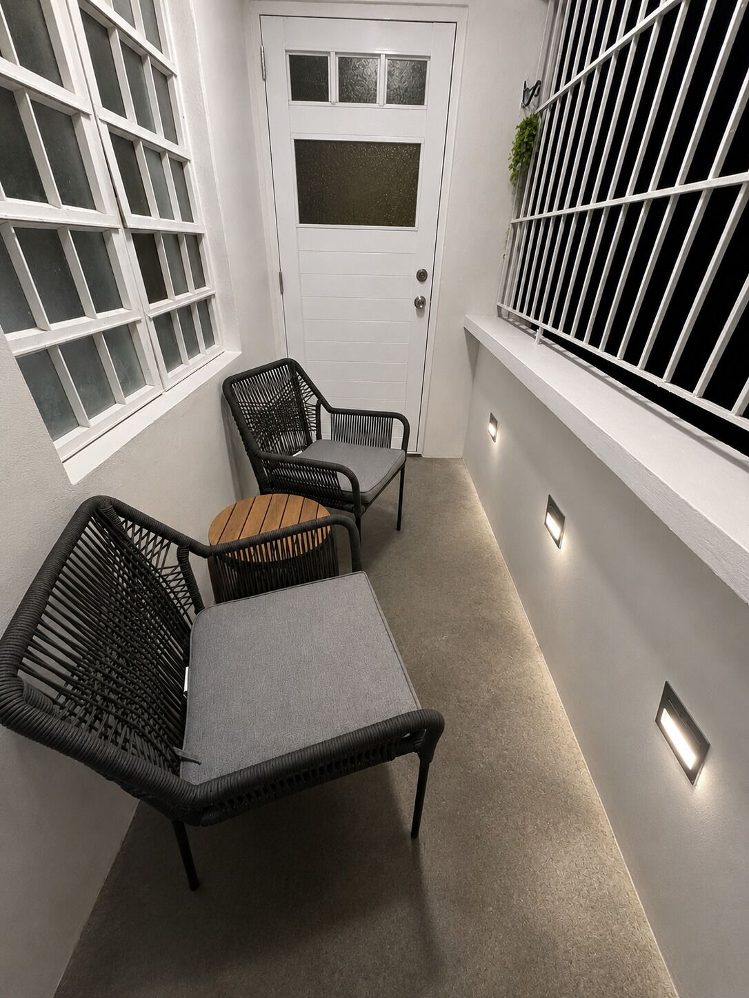 A small, enclosed outdoor space with a white door, two wicker chairs, a small table, and a window with a metal grill.