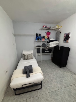 A small, single-bed room with a metal frame bed, a white mattress, and a grey blanket. There is a shelf with folded clothes and a black bag.