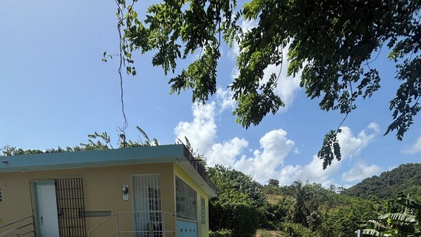 A building with a staircase leading to a door, a fenced area with grass, and a clear sky. - The Naguabeña House of the Anvil (Ceiba)
