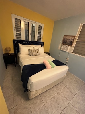 A bed with a headboard, pillows, and a blanket. There is a nightstand with a lamp and a picture frame on the wall.