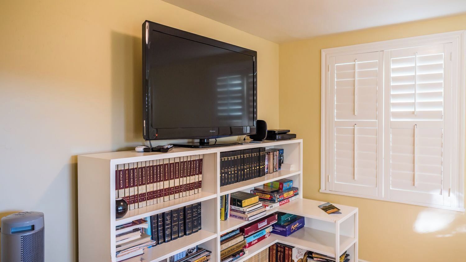 A room with a television on a stand, a bookshelf filled with books, and a window with blinds.