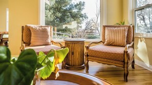 Ein Zimmer mit zwei Polstersesseln, Kissen mit Muster, einem runden Holztisch und Blick auf Bäume und einen Zaun durch ein Fenster.