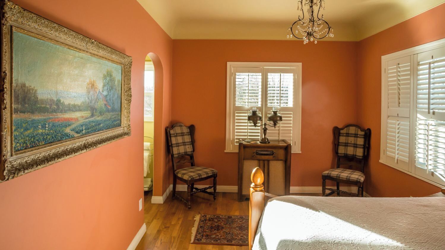 Um quarto com uma cama, duas cadeiras, uma mesa de cabeceira, um quadro e uma janela com persianas.