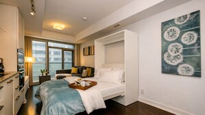 Une chambre d’hôtel comprenant un lit, une table de chevet avec un plateau, une fenêtre avec des rideaux, un tableau au mur et une kitchenette.