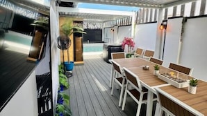 An outdoor patio with a wooden table and chairs, surrounded by potted plants and a metal ceiling structure.