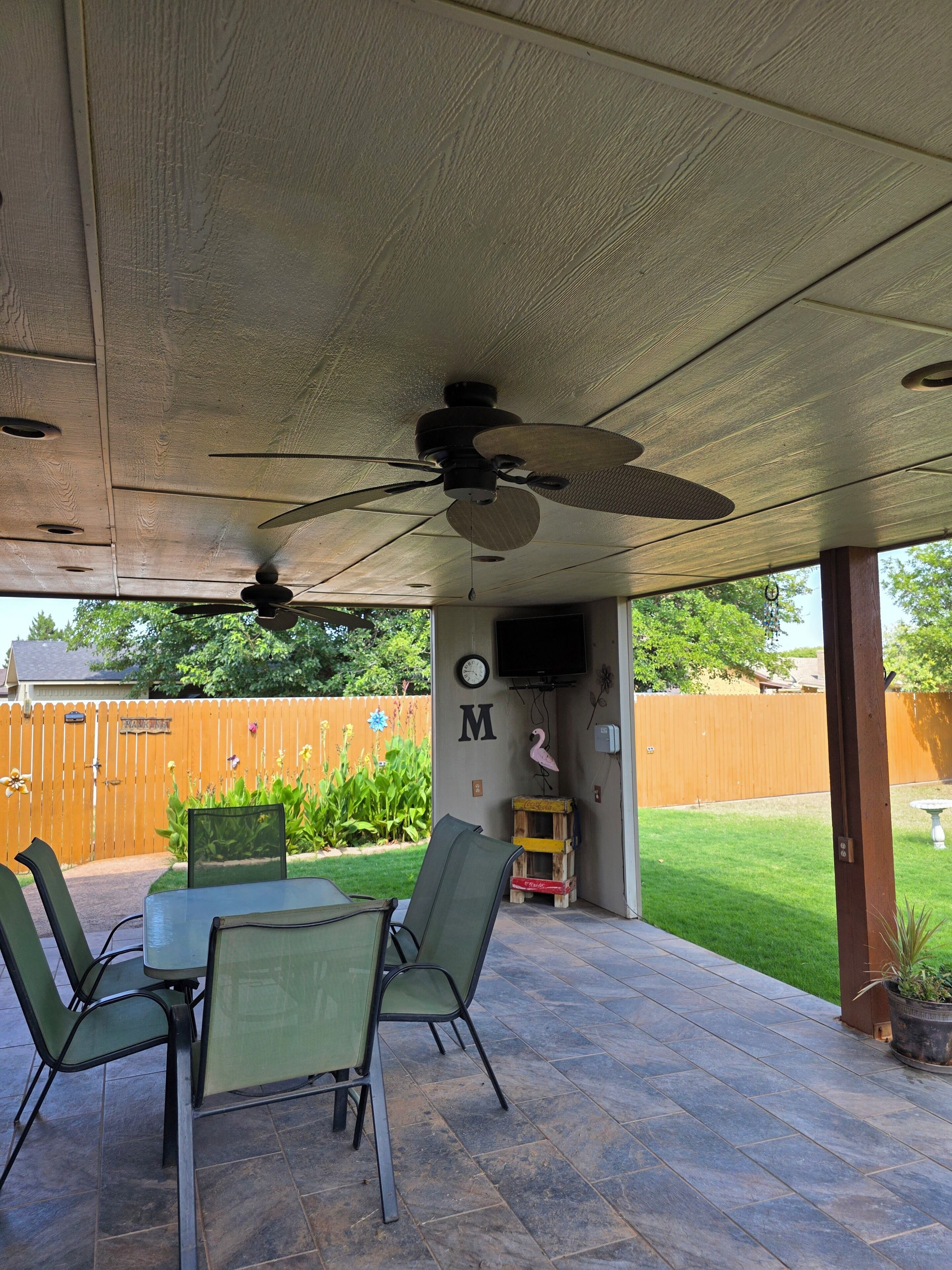 An outdoor patio with a table and chairs, a ceiling fan, and a wooden fence in the background.