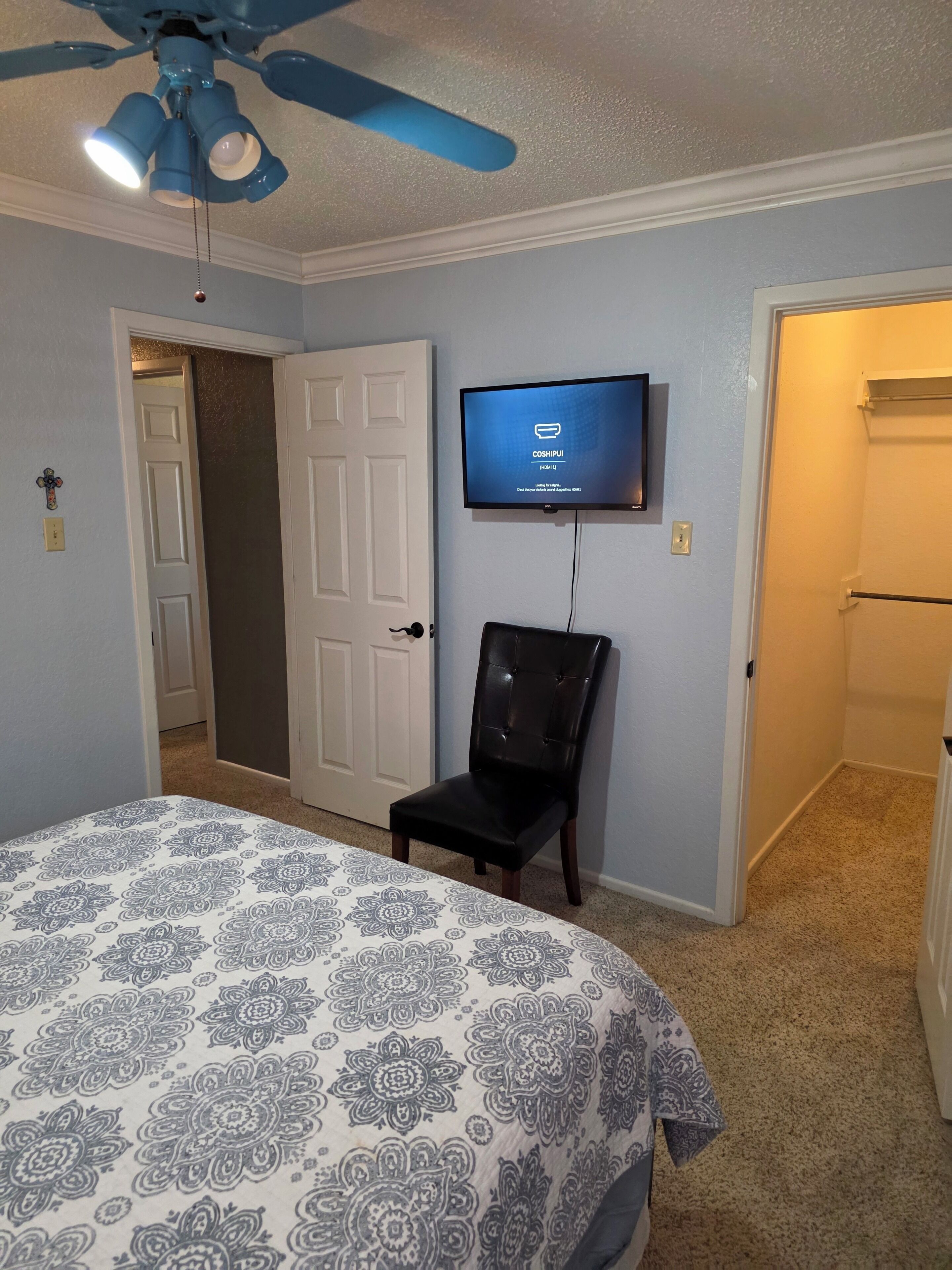 Un dormitorio con cama, televisor montado en la pared, ventilador de techo y puerta que da a otra habitación.