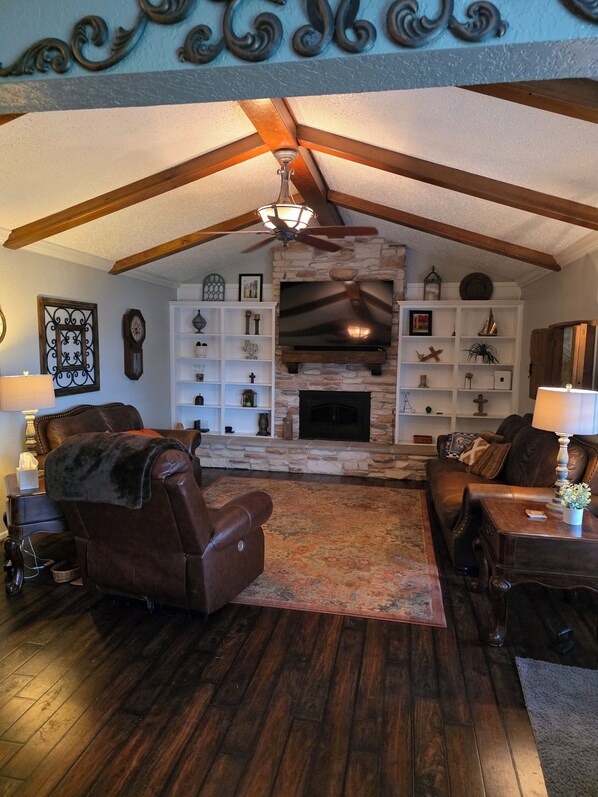 A cozy living room with a fireplace, wooden beams, and a ceiling fan.