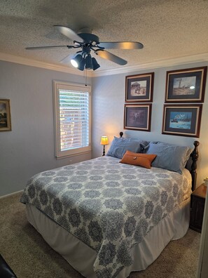 A bedroom with a bed, ceiling fan, window with blinds, and framed pictures on the wall.