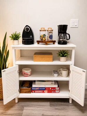 Uma estante branca aberta com uma cafeteira, potes e uma planta na prateleira superior, e uma caixa e canecas de café na prateleira do meio.