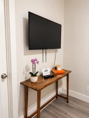 Un televisor de pantalla plana montado en la pared sobre un mueble bajo de madera, con una planta en maceta, un letrero de Wi-Fi y un control remoto.