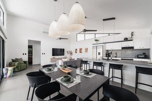 A modern kitchen with white cabinets, a central island, and a dining area with a dark table and black chairs. - Modern Villa | Private Pool | Near Playa Grande (Matapalo)