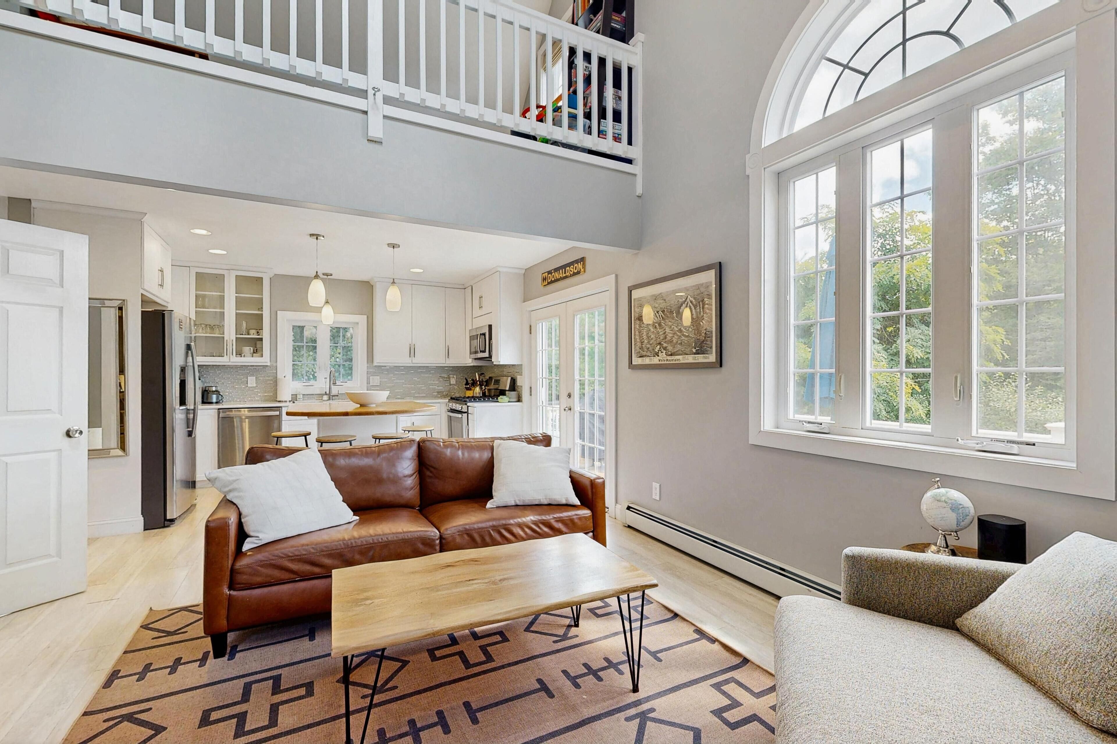 A spacious living area with a brown leather sofa, a wooden coffee table, and a view of the kitchen and upper level.