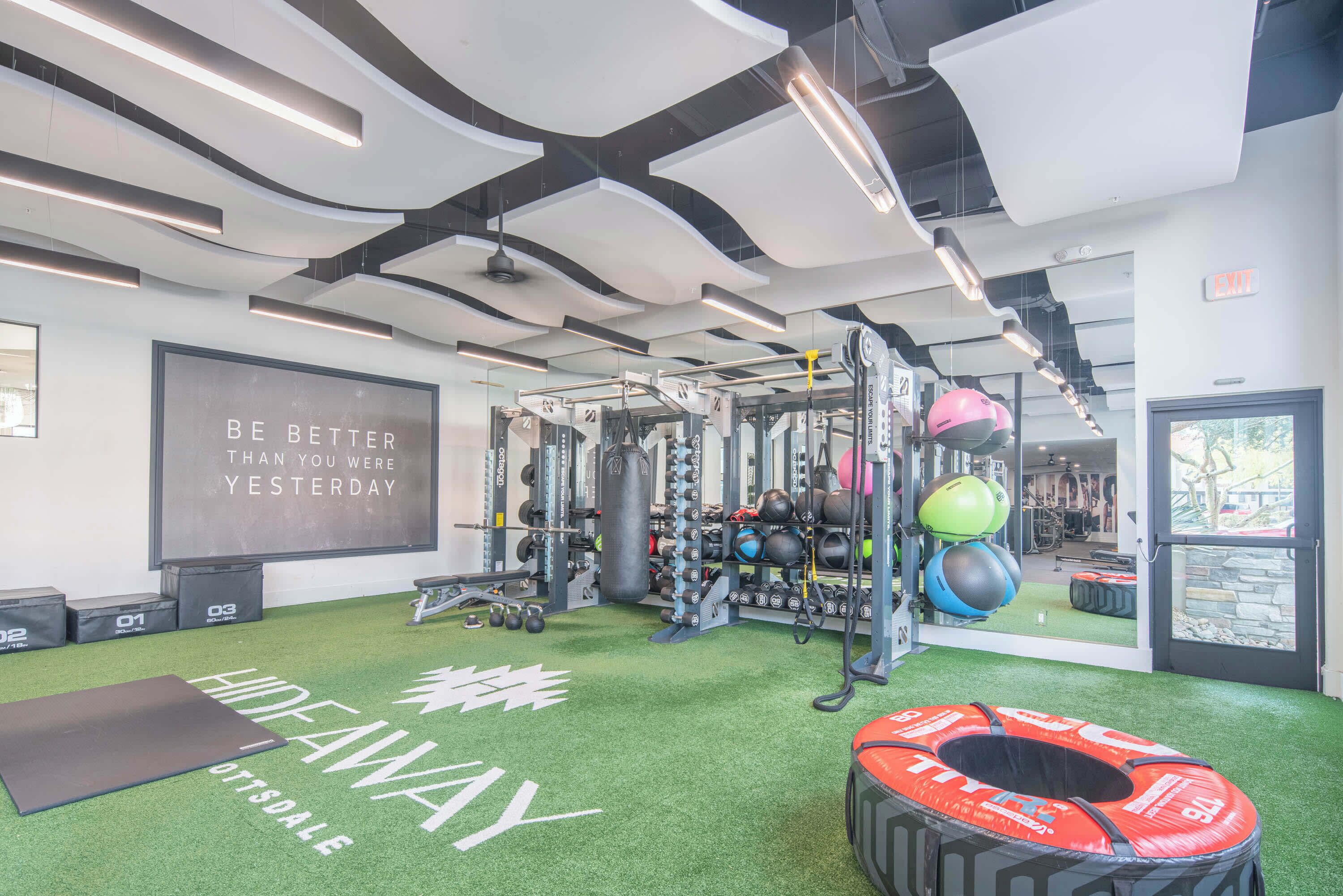 Un gimnasio con césped artificial verde, equipamiento de levantamiento de pesas y una frase motivacional en la pared.