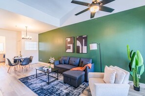 A modern living room with a green accent wall, a ceiling fan, a sofa, a coffee table, and a dining area with chairs. - Steps to Kierland Balcony Pool Parking WD WiFi (Scottsdale)