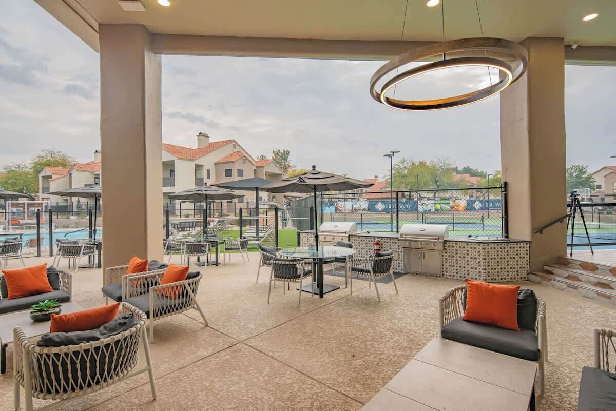 Un patio exterior con mobiliario de mimbre, una parrilla y vistas a una piscina y edificios al fondo.